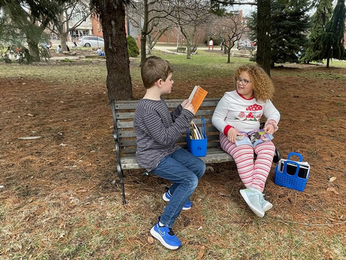A student reads to another student on a bench outside.